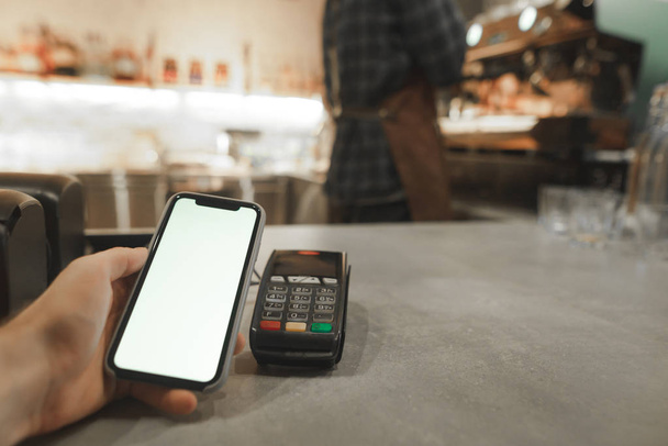Mobile payment in cafe with smartphone nfc near field communication wireless technology. Paying with smartphone at the coffee shop, while male barista is preparing coffee at the coffee machine. - Foto, Bild