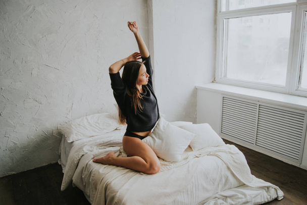 Beautiful tender girl in black lingerie and jumper is sitting on the bed and stretching herself. A young woman basks in bed with white linens. The girl woke up and enjoys a good morning. - Fotoğraf, Görsel