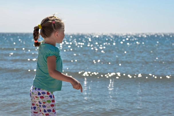Little pretty girl on summer beach. Cheerful little girl is looking at blue sky and summer sea. Happy child girl playing at beach and having fun - Фото, зображення