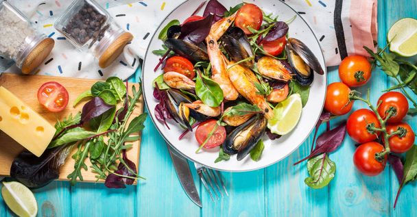 Fresh seafood salad, mussels, shrimp, fresh vegetables and herbs on blue wooden table. Top view - Photo, Image