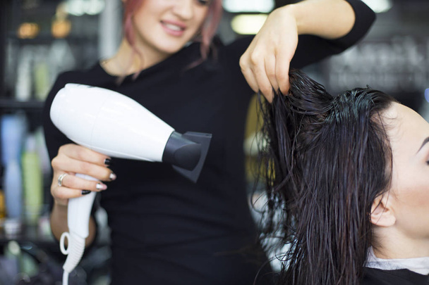 Профессиональный парикмахер сушит волосы красивой молодой женщины в салоне красоты. Профессиональный уход
 - Фото, изображение