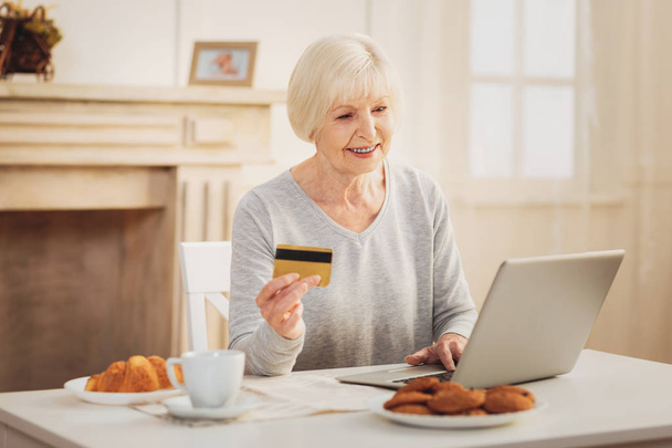 Shopping online. Modern smiling pensioner feeling busy while shopping online having tasty French breakfast - 写真・画像