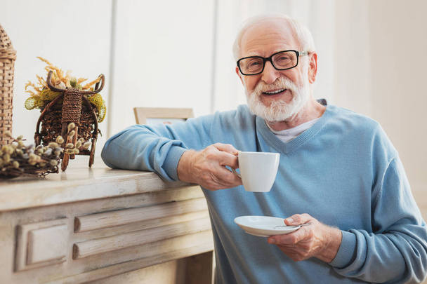 Home fireplace. Respectful elderly man feeling relaxed while drinking tasty tea standing near fireplace at home - Фото, изображение