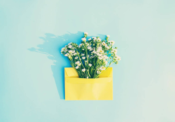 Gele papieren envelop met kleine tuin witte kamille bloemen op lichtblauwe achtergrond. Feestelijke bloemen sjabloon. Wenskaart ontwerp. Bovenaanzicht. Vintage stijl. - Foto, afbeelding