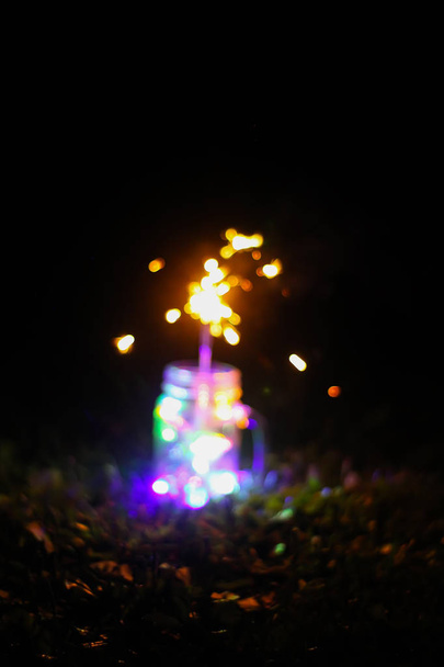 Świąteczna kartka z jasnymi kolorowymi lampkami girlandowymi i bengalskim ogniem w szklanym słoiku na nocnym tle natury. - Zdjęcie, obraz
