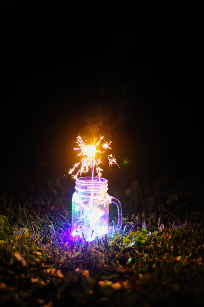 Праздничная открытка с яркими красочными гирляндами и бенгальским огнем в стеклянной банке на ночном фоне природы. - Фото, изображение