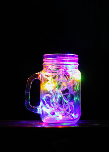Gece karanlığında cam kavanozda parlak renkli çelenk ışıkları olan bir kart.. - Fotoğraf, Görsel