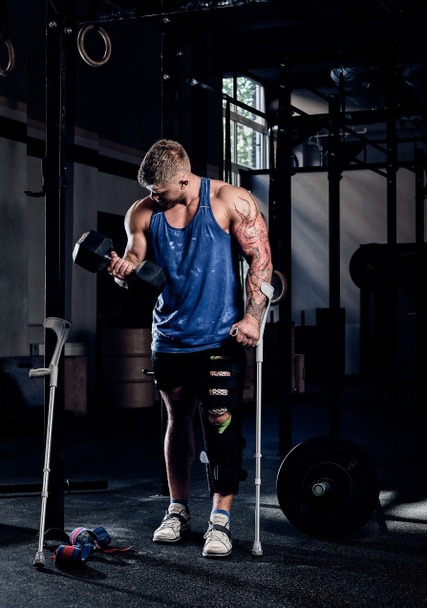 Professional bodybuilder with a tattoo on his hand, warming up on a gym, doing exercises with a dumbbell while leaning on a crutch. - Foto, Bild