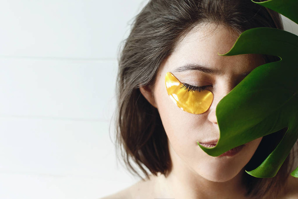 目の皮膚のケアと治療。ナチュラルメイクと緑のモンステラの葉に黄金の目パッチと美しい若い女性の肖像画。自然のスキンケア。目の前でリフティングの抗しわコラーゲン パッチ - 写真・画像