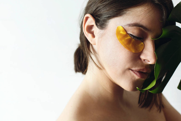 Lifting anti-wrinkle collagen patches under eyes. Eye Skin Care. Portrait of beautiful young woman with golden eye patches at green monstera leaf on white background, copy space. Natural skin care. - Fotoğraf, Görsel