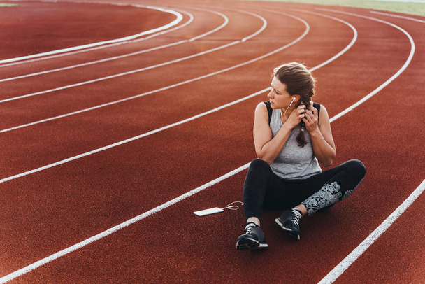 A young beautiful woman having rest with her smartphone sitting at a stadium running track. - Foto, imagen