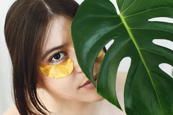 Portrait of beautiful young woman with natural makeup and golden eye patches at green monstera leaf. Natural skin care. Lifting anti-wrinkle collagen patches under eyes. Eye Skin Care and Treatment. - Photo, Image
