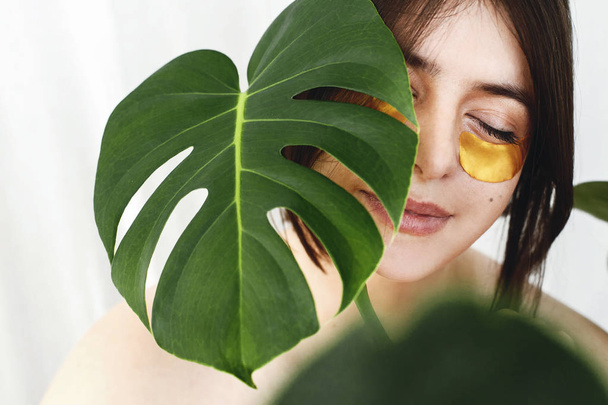 Portrait of beautiful young woman with natural makeup and golden eye patches at green monstera leaf on white background. Natural care. Lifting anti-wrinkle collagen patches under eyes. Eye Skin Care - Фото, зображення