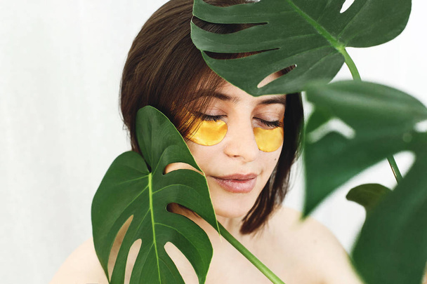 Ευτυχισμένος κορίτσι με φυσικό δέρμα και ανύψωσης μπαλώματα κολλαγόνου κατά των ρυτίδων κάτω από τα μάτια και πράσινο palm leaf. Πορτρέτο του όμορφη νεαρή γυναίκα με τα χρυσά μάτια μπαλώματα με λευκό. Φροντίδα του δέρματος μάτι - Φωτογραφία, εικόνα