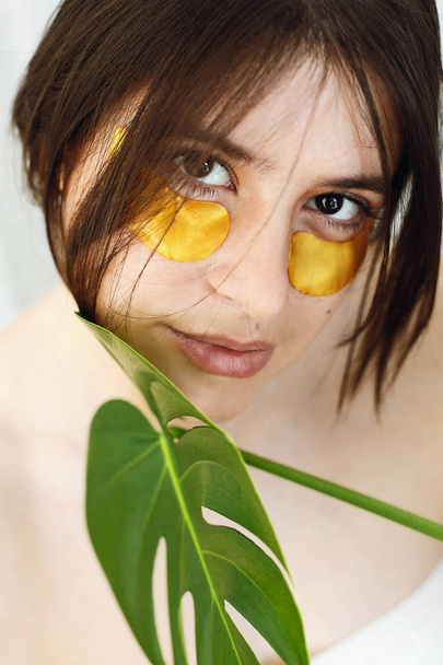 Šťastná dívka s přírodní kůží a zvedací Protivrásková kolagenu náplasti pod oči a zelené Palmový list. Portrét krásné mladé ženy s Zlaté oko skvrny na bílém pozadí. Oční péče o pleť - Fotografie, Obrázek
