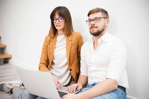 Два молодых сотрудника одеты непринужденно, используя ноутбук в белом интерьере
 - Фото, изображение