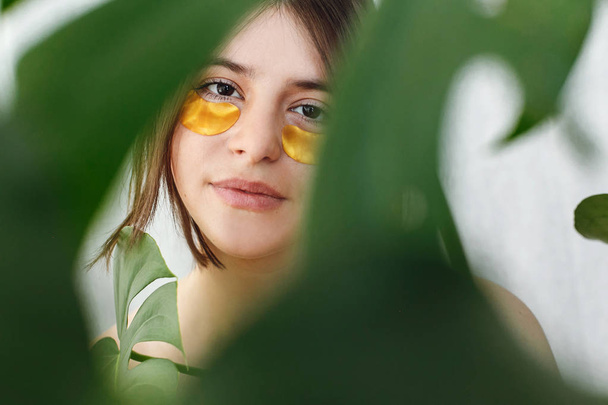 Portret van mooie jonge vrouw met natuurlijke make-up en gouden oog patches op groene monstera blad. Natuurlijke huidverzorging. Opheffing anti-rimpel collageen patches onder ogen. De zorg van de huid van het oog en behandeling. - Foto, afbeelding