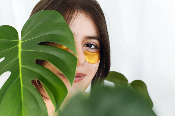Happy girl with natural skin and lifting anti-wrinkle collagen patches under eyes and green palm leaf. Portrait of beautiful young woman with golden eye patches on white background. Eye Skin Care - Photo, Image