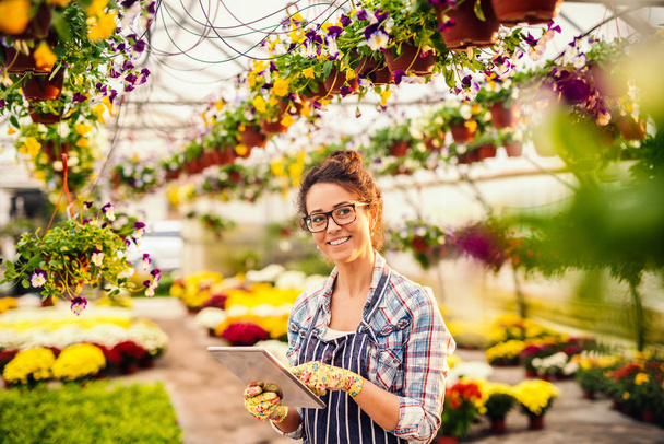 Portrait of beautiful florist with eyeglasses, brown hair and apron while standing in greenhouse. All around flowers. - Valokuva, kuva