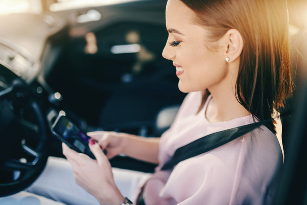Piękny Kaukaska brunetka siedzi w samochodzie i pozwania inteligentny telefon do czytania lub wysłanie wiadomości. - Zdjęcie, obraz