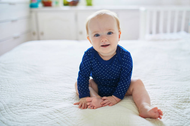 Baby girl sitting on bed and laughing. Happy healthy little child in blue clothes. Infant kid in sunny nursery - Foto, Imagem