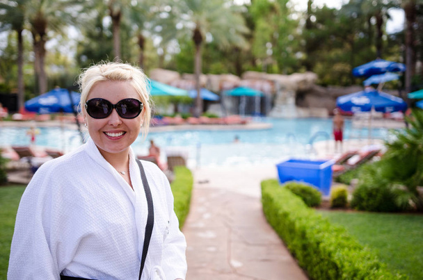 Mujer rubia vestida con una bata de spa blanca en una zona de piscina al aire libre de un resort de lujo
 - Foto, Imagen