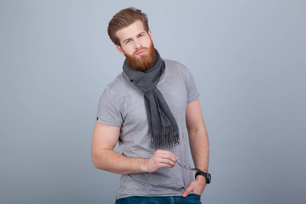 young handsome bearded man standing by the grey wall wearing scarf and grey t-shirt and holding sunglasses in his hand looking straight to the camera. - Valokuva, kuva