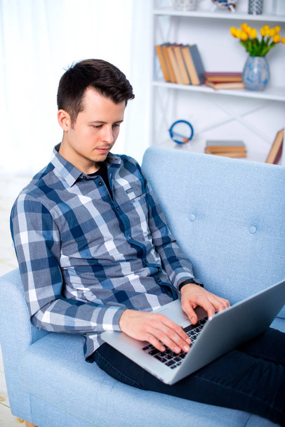 Nuori viehättävä kaveri mies on selaamassa hänen kannettava tietokone, istuu kotona kodikas sininen sohva kotona, yllään rento asu
. - Valokuva, kuva