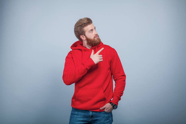 fiatal jóképű szakállas ember visel piros pulóver állandó, a szürke háttér, tartja a kezét a zsebében, és mutatva az ujját a jobb oldalon, és ott a érdeklődéssel. - Fotó, kép