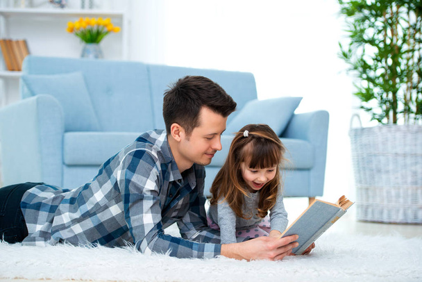 Cute little girl listening to dad reading fairy tale lying on warm floor together, caring father holding book , family hobbies activities at home. - Zdjęcie, obraz
