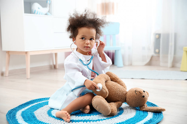 Cute African American child imagining herself as doctor while playing with stethoscope and toy bunny at home - Фото, изображение