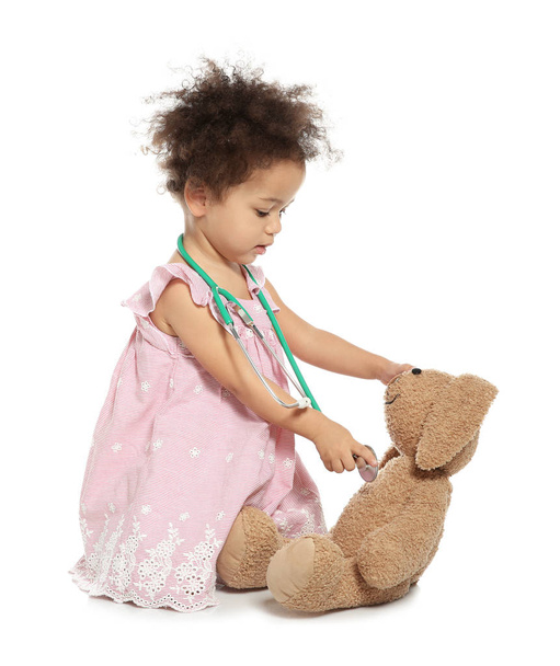 Cute African American child imagining herself as doctor while playing with stethoscope and toy bunny on white background - Zdjęcie, obraz