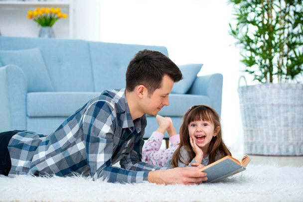 Cute little girl listening to dad reading fairy tale lying on warm floor together, caring father holding book , family hobbies activities at home. - Фото, изображение