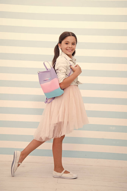 Simply cuteness. Child girl cute smiling face carry bag on shoulder. Kid long curly hair wear fashionable outfit. Girl with cute bag walks striped background. She likes try on fashionable outfits - Foto, Imagen