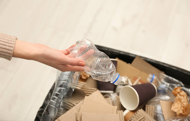 Young woman throwing plastic bottle in trash bin indoors, closeup. Waste recycling - Фото, изображение