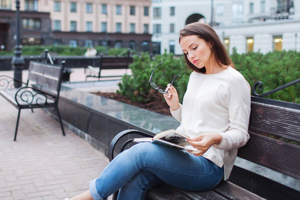 A beautiful young girl with long brown hair sitting on a bench with a book, holding eyeglasses. She left the house on a warm evening to read in the yard. The urban background. - Foto, Bild