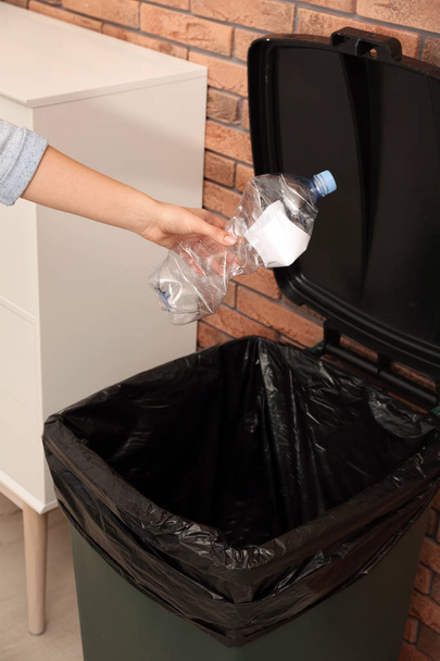 Young woman throwing empty plastic bottle in trash bin indoors, closeup. Waste recycling - Fotoğraf, Görsel