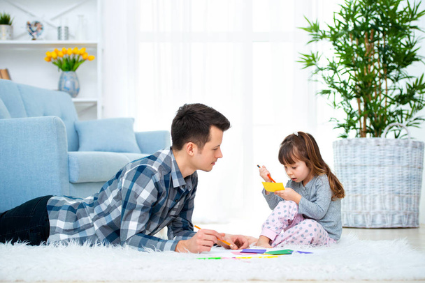 Vater und kleine Tochter haben gute Familienzeit zu Hause. Papa mit Mädchen auf warmem Boden liegend Zeichnung mit bunten Filzstiften. - Foto, Bild