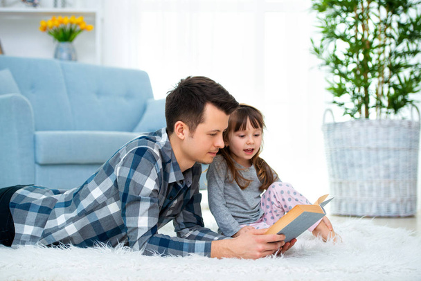 Cute little girl listening to dad reading fairy tale lying on warm floor together, caring father holding book , family hobbies activities at home. - Foto, immagini