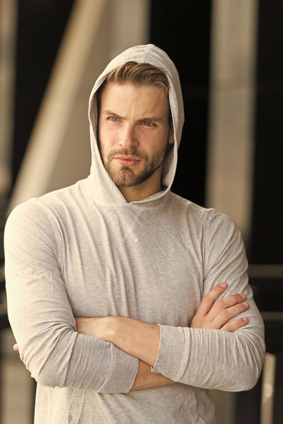 Cool and confident. Man unshaven hooded folded arms looks handsome and cool. Guy bearded attractive cares about appearance. Man bristle serious face, urban background, defocused. Metrosexual concept - Photo, Image