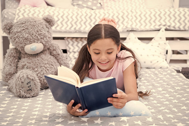 Read fairytale in bed. Girl child lay bed with teddy bear read book. Kid prepare to go to bed. Time for evening fairytale. Girl kid long hair cute pajamas relax and read fairytale book to bear toy - Foto, imagen
