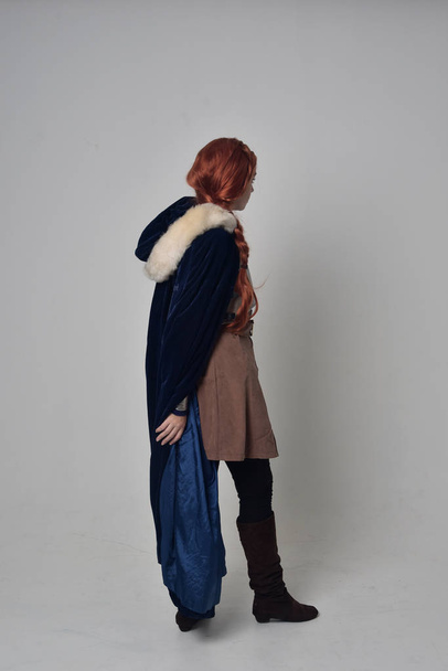 ολόσωμο πορτρέτο του ένα κόκκινο πλασματάκια κορίτσι φορώντας κοστούμι μεσαιωνικός πολεμιστής, μπλε βελούδο μανδύα και χαλύβδινη θωράκιση, στέκεται πόζα σε φόντο γκρι στούντιο. - Φωτογραφία, εικόνα