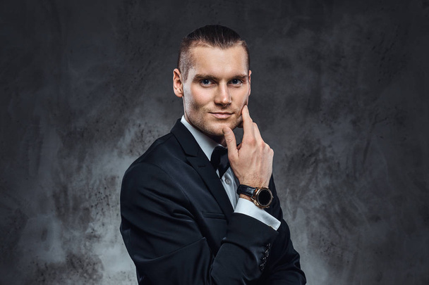 Elegantly dressed handsome man wearing a black classical suit and bow tie, touching his face and looking at a camera. Studio shot on a dark textured wall - Zdjęcie, obraz