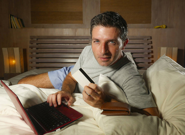 young attractive and relaxed man at home bedroom lying on bed buying or banking online using credit card for paying in electronic business and commerce concept - Photo, Image