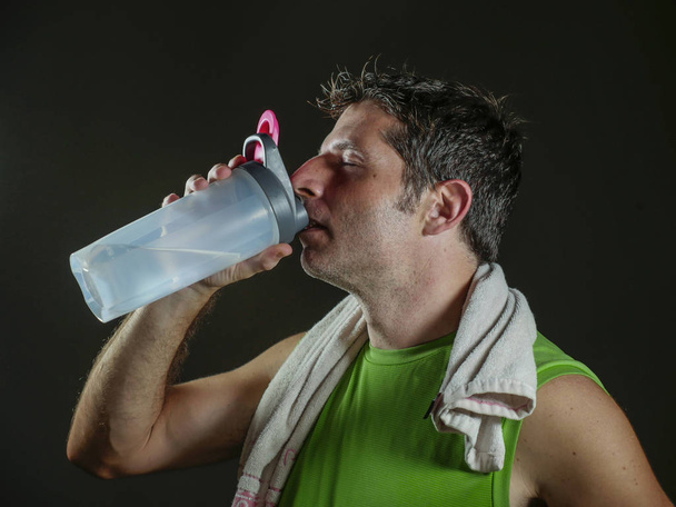 young sweaty attractive and fit sport man drinking water holding bottle cooling off after hard fitness workout at gym club isolated on black background in healthy lifestyle and training - Foto, Imagen