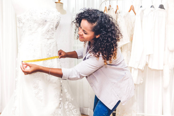 lächelnde afrikanisch-amerikanische schwarze Modedesignerin, die im Atelier der Werkstatt steht und mit Klebeband am Kleid der Schaufensterpuppe sitzt - Foto, Bild