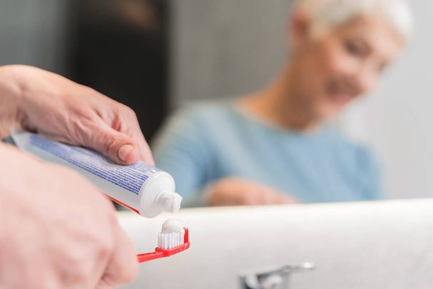 senior woman putting tooth paste on toothbrush in her bathroom  - Zdjęcie, obraz