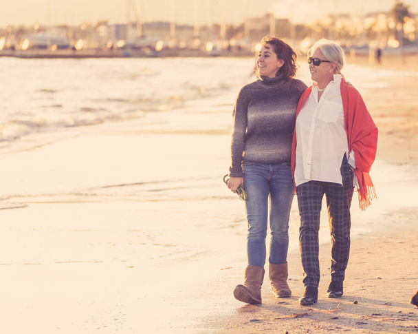 Portrait of happy senior mother and adult daughter spending time together holding hands laughing and walking on beach at sunset light in Happy family moments Generations Retirement and People concept. - Фото, изображение