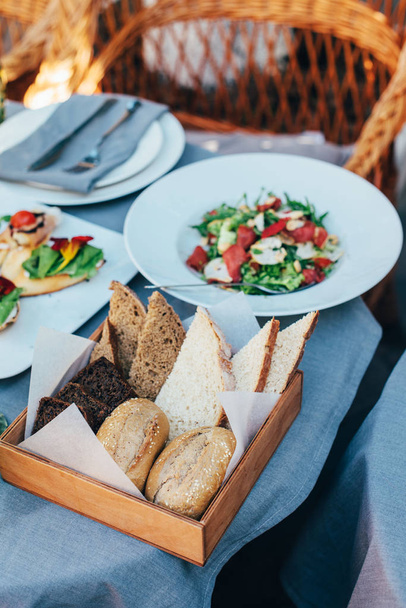 Fresh Italian style salad, served in a white plate, and a basket with bread is located next to it - Fotó, kép