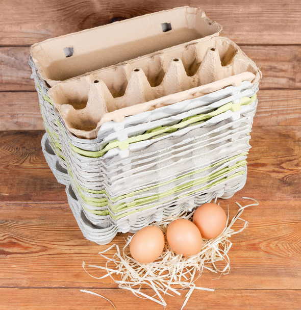 Стек різних відкритих порожніх яєчних коробки з переробленої паперової целюлози і кілька коричневих курячих яєць поруч з ними на старому дерев'яному сільському столі
 - Фото, зображення